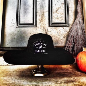 May include: A black sun hat with a wide brim, featuring a patch that reads "ENTERING EST. 1626 SALEM" with a rabbit graphic. The hat is displayed on a decorative stand, with a broom and pumpkin in the background.