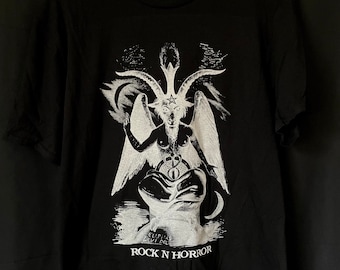 May include: Black t-shirt featuring a white graphic of a horned figure with wings, a pentagram, and a crescent moon. The text "ROCK N HORROR" is printed below the graphic. The shirt is hanging on a wooden hanger.