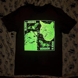 Könnte beinhalten: Schwarzes T-Shirt mit einem grün leuchtenden Grafikdruck von zwei Katzen mit Fledermäusen und dem Text "Bloody Hisses" und "Rock 'n Horror".