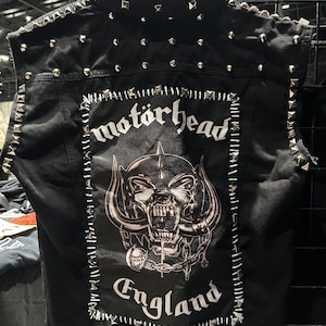 May include: Black denim vest adorned with silver pyramid studs on the shoulders and edges. The back displays a large graphic of a skull with horns, chains, and the band name "Motörhead" and the word "England" in white.