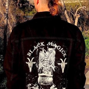 Könnte beinhalten: Eine schwarze Jeansjacke mit einem weißen Grafikdruck auf der Rückseite. Die Grafik zeigt einen geflügelten Dämon, der einen Kessel hält, mit dem Text "Black Magick 1692 The Devil is in Salem Rock N Horror".