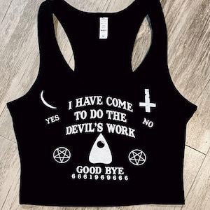 Puede incluir: Top corto negro con diseño de espalda nadadora. La camiseta presenta texto y gráficos blancos, incluyendo una luna creciente, una cruz, pentagramas y la frase "I HAVE COME TO DO THE DEVIL'S WORK". La parte inferior de la camiseta dice "GOOD BYE" y una serie de números.