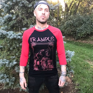 May include: A black and red baseball tee shirt with a graphic of a creature with horns and the word "Krampus" printed in pink.