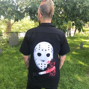 May include: Black short-sleeved shirt featuring a white hockey mask with black eye holes and a red blood splatter design. The shirt is worn by a person standing outside on a sunny day.