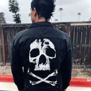 May include: Black denim jacket with a white skull and crossbones design on the back. The skull has a lightning bolt through it.