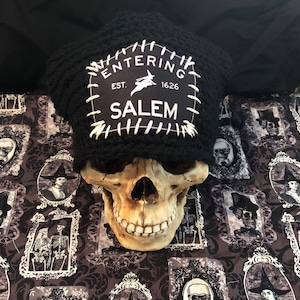 May include: A skull wearing a black knit hat with a patch that reads "ENTERING SALEM EST. 1626" with a witch graphic. The skull rests on a patterned fabric with framed portraits of witches and skeletons.