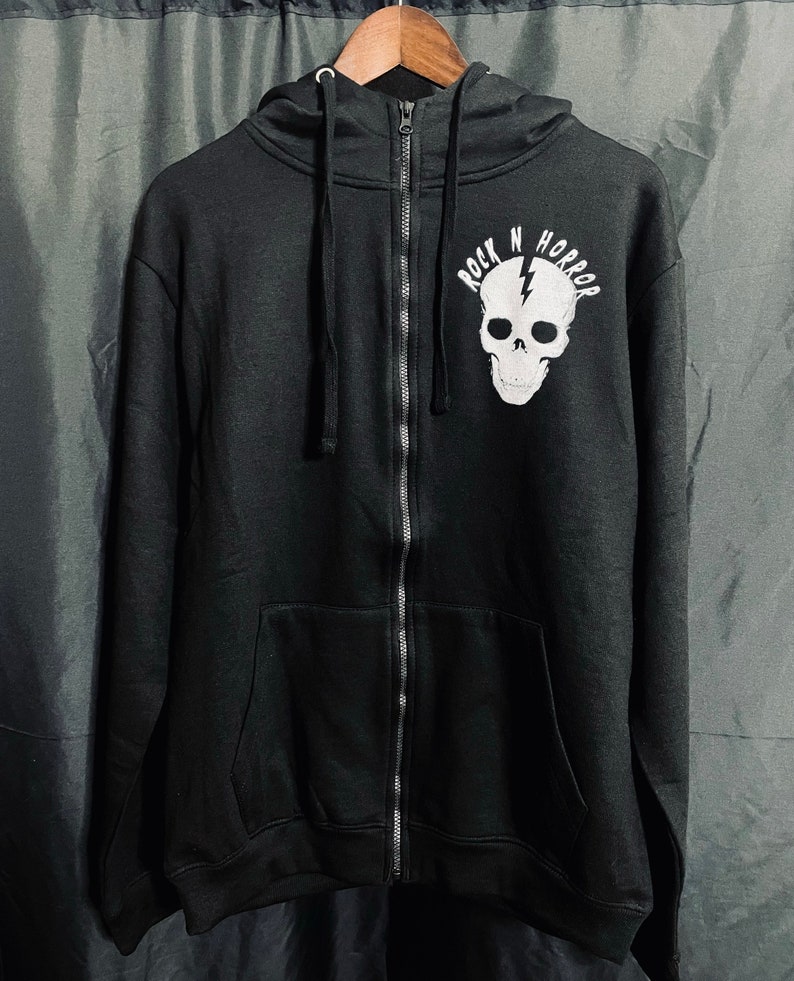 Puede incluir: Sudadera negra con cremallera y un gr&aacute;fico de calavera blanca y rayo. El texto "ROCK N HORROR" est&aacute; encima de la calavera. La sudadera con capucha tiene una capucha con cord&oacute;n y un bolsillo delantero. Cuelga de una percha de madera.