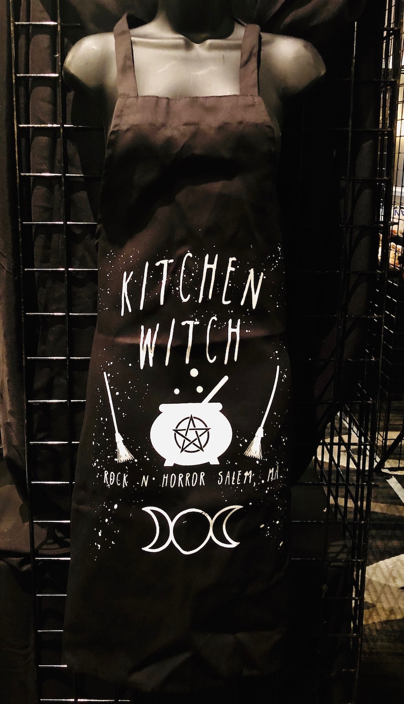 May include: Black kitchen apron with white text that reads "Kitchen Witch" and a cauldron with a pentagram inside. The apron also has a broom, moon phases, and the text "Rock N Horror Salem, MA".