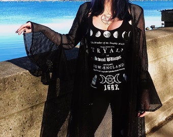 May include: A woman in a black hat, a black mesh duster, and a black tank top with a pentagram necklace. The tank top features the text "TRYAL of Several Witches, NEW-ENGLAND 1692". The background shows a body of water.