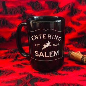 May include: Black ceramic mug with a white rectangular label featuring the text "ENTERING SALEM" and a silhouette of a witch on a broom. The mug has a black handle and is set against a red and black patterned background.
