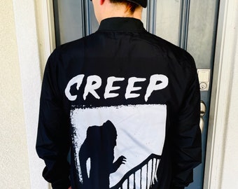 May include: Black bomber jacket with the word "CREEP" in white block letters across the upper back. Below the word is a white graphic of a shadowy figure descending a staircase. The jacket has a black ribbed collar, cuffs, and hem.