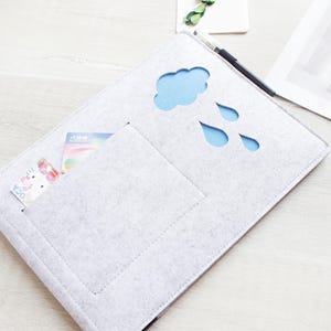 May include: A light gray felt laptop sleeve with a cloud and raindrop cutouts revealing a blue interior. A pocket holds a card. A pen and magazine are nearby.