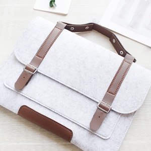 May include: A light gray felt laptop bag with brown leather straps and accents. The bag has a flap closure with two adjustable buckles and a handle. The bag is sitting on a light wood surface.