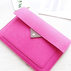 May include: A bright pink felt envelope-style clutch with a flap closure. The clutch features a small, gray leather accent with a silver button. A white notepad with green accents and a magazine are in the background.