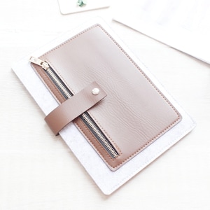 May include: A brown leather tablet case with a zipper closure and a brown strap secured by a silver button. The case is on top of a light gray fabric, with a white background.