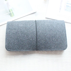 Puede incluir: Una cartera o estuche organizador rectangular de fieltro gris oscuro con esquinas redondeadas y una banda elástica negra fina envuelta verticalmente alrededor del centro para asegurar el cierre. El material tiene una textura fibrosa.