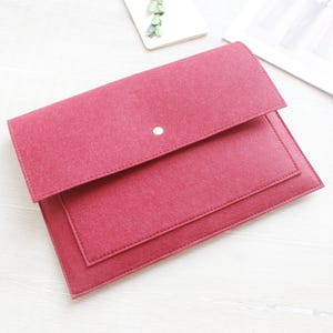 May include: A rectangular felt laptop sleeve in a deep magenta colour. The sleeve features a fold-over flap secured by a single silver-toned metal snap button closure. The material has a visible textured finish with stitched edges along the perimeter.