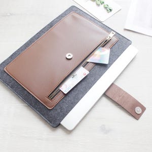 May include: A brown leather laptop sleeve with a zippered pocket and a gray felt base. The sleeve has a magnetic snap closure and a brown leather strap. A laptop is partially inserted into the sleeve.