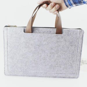 May include: A light gray felt laptop sleeve featuring a rectangular shape with rounded corners and two brown leather handles attached to the top. The sleeve includes a black zipper closure along the top edge and a stitched border detail on the front.