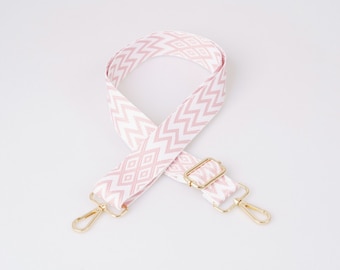 May include: A white and pink patterned adjustable strap with gold metal hardware. The strap is made of a woven fabric with a geometric design.