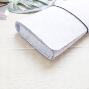 May include: A light gray felt sleeve with rounded corners and a thin black elastic band wrapped around the exterior to secure the closure. The material has a soft, textured appearance and the case is rectangular in shape with a flat profile.
