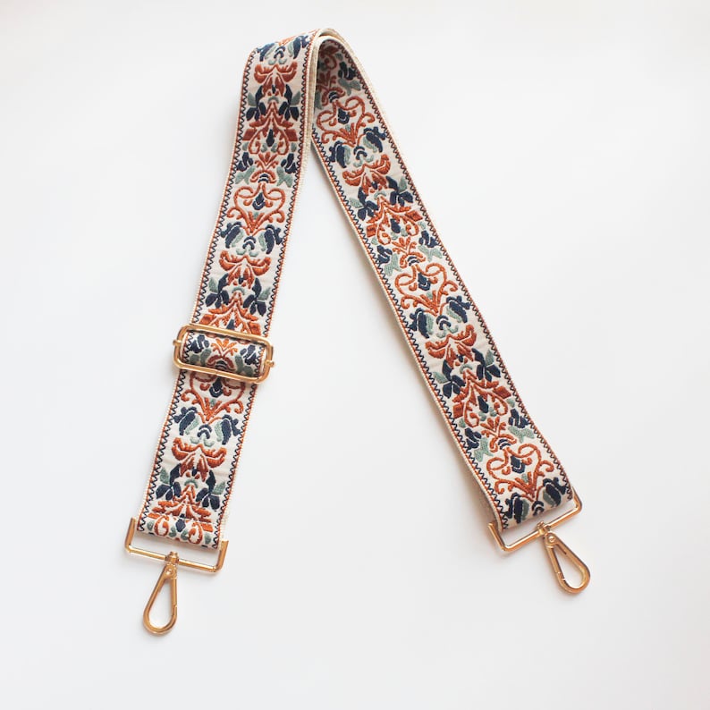 May include: A white woven strap with a gold buckle and clasps. The strap has a floral pattern in shades of orange, blue, and green.