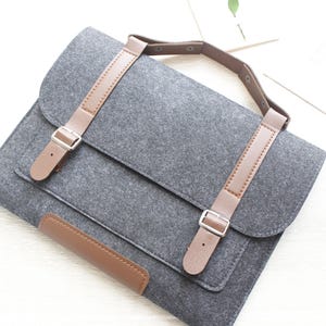 Puede incluir: Bolso gris de fieltro para portátil con correas y detalles de cuero marrón. El bolso tiene un cierre de solapa con dos correas ajustables y un asa. Diseñado para llevar un portátil o una tableta, tiene un diseño minimalista.