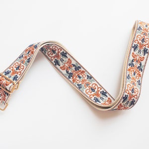 May include: A white, blue, and orange patterned fabric strap with a gold metal clasp. The strap is designed for a bag or purse.