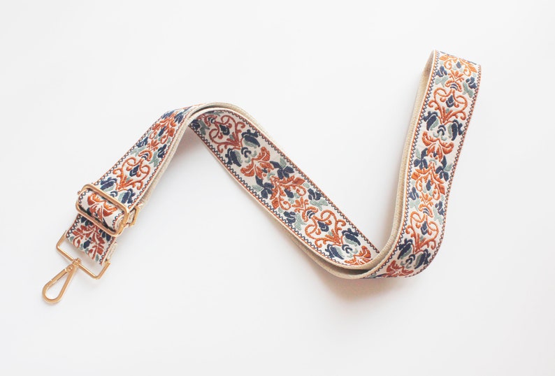 May include: A white, blue, and orange patterned fabric strap with a gold metal clasp. The strap is designed for a bag or purse.