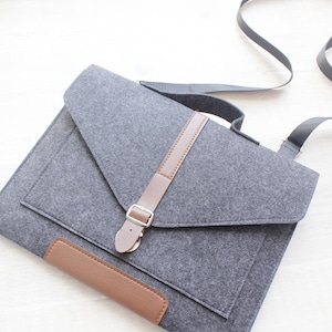 May include: Gray felt messenger bag with a brown leather accent on the flap and a matching leather strap closure. The bag has a long, adjustable black strap for shoulder or crossbody wear.