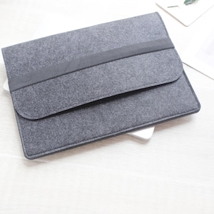 Könnte beinhalten: Eine graue Filz-Laptoptasche mit einem dunkelgrauen Bandverschluss. Die Tasche ist rechteckig mit einer abgerundeten Klappe. Das Produkt befindet sich auf einer weißen Oberfläche.