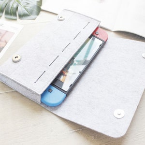 May include: A gray felt case with a zipper closure and snap buttons. The case is open and a Nintendo Switch console is inside.
