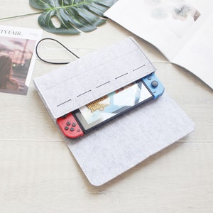Puede incluir: Una funda de transporte de fieltro gris con una correa negra para una consola Nintendo Switch. La consola está dentro de la funda con los Joy-Cons azules y rojos conectados.