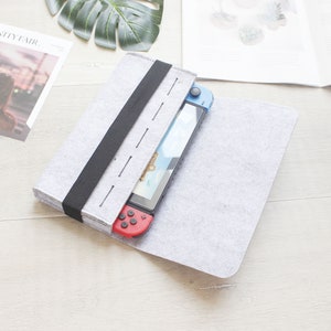 May include: A gray felt case with a black strap and a Nintendo Switch console inside. The console has red and blue buttons and a blue screen.