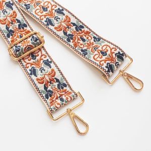 May include: A white fabric strap with a gold metal clasp and a floral pattern in shades of orange, blue, and green.