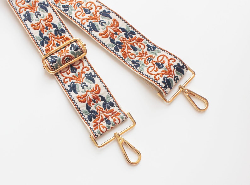 May include: A white fabric strap with a gold metal clasp and a floral pattern in shades of orange, blue, and green.