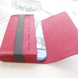 May include: A burgundy felt case with a black elastic band, designed to hold a handheld gaming device. The case is open, revealing the device inside. The case is on a white surface.