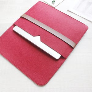 May include: A red felt laptop sleeve with a brown leather strap closure. The sleeve holds a silver laptop, and is open to display the device. The sleeve is on a white surface.