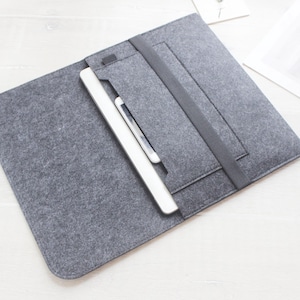May include: Grey felt laptop sleeve with a black elastic band closure. A white tablet and stylus are inserted into the sleeve's pocket. The sleeve is designed to protect and carry electronic devices.