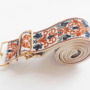 May include: A white and beige woven strap with a gold metal buckle and clasp. The strap has a floral pattern in shades of orange, blue, and green.
