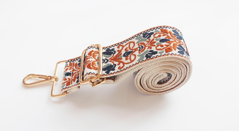 May include: A white and beige woven strap with a gold metal buckle and clasp. The strap has a floral pattern in shades of orange, blue, and green.
