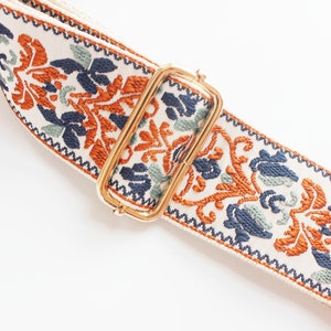 May include: A white fabric strap with a gold metal buckle. The strap has a colorful floral pattern in shades of orange, blue, and green.