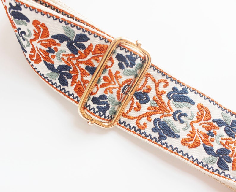 May include: A white fabric strap with a gold metal buckle. The strap has a colorful floral pattern in shades of orange, blue, and green.