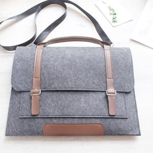 Può includere: Borsa messenger in feltro grigio con dettagli in pelle marrone, tra cui cinghie, fibbie e manico. La borsa ha una chiusura a patta e una tracolla. Il design è semplice e moderno, adatta per trasportare un laptop o altri oggetti.