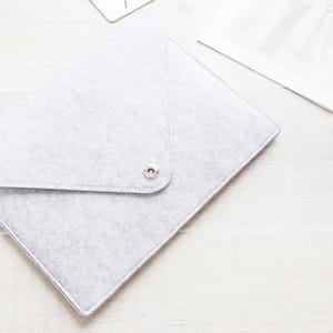 May include: A light grey felt laptop sleeve with an envelope-style flap closure. The sleeve features a single silver-toned metal snap button centred on the triangular flap. The material has a soft, textured appearance with visible stitching.