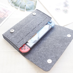 May include: A gray felt case with a zipper closure and a Nintendo Switch console inside. The case has a soft, textured feel and is designed to protect the console from scratches and bumps.