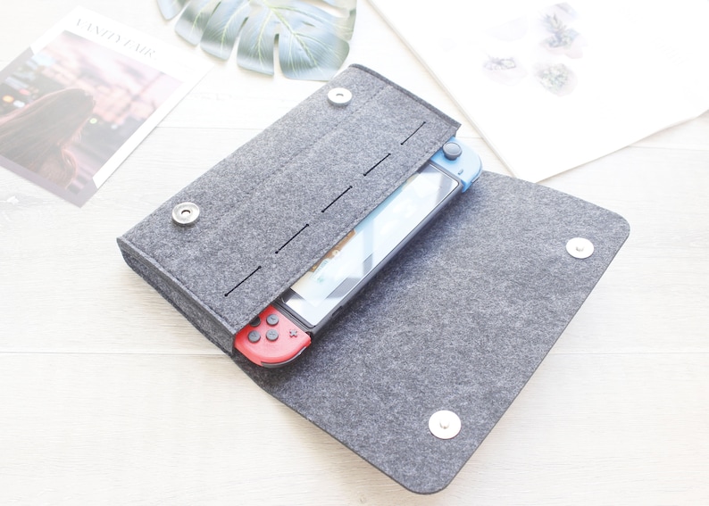 Pode incluir: Uma capa de feltro cinza com fecho magn&eacute;tico, contendo um console Nintendo Switch com controles vermelho e azul. A capa est&aacute; aberta, revelando a tela. A capa foi projetada para proteger o console.