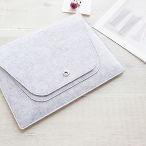 May include: A light gray felt laptop sleeve with a flap closure and a silver snap. The sleeve is rectangular with rounded corners and a stitched edge. A magazine and a small flower are in the background.