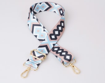 May include: A colorful patterned strap with gold hardware. The strap is made of a woven fabric with a geometric design in shades of blue, pink, and black. The strap has two gold clasps.