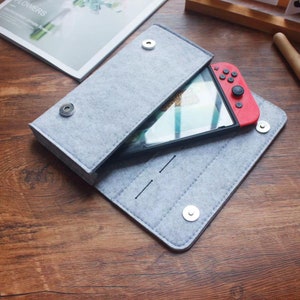 Op de afbeelding: Een grijze wollen etui met een ritssluiting en een drukknoop. De etui is open en een rood-zwarte Nintendo Switch-console bevindt zich erin.